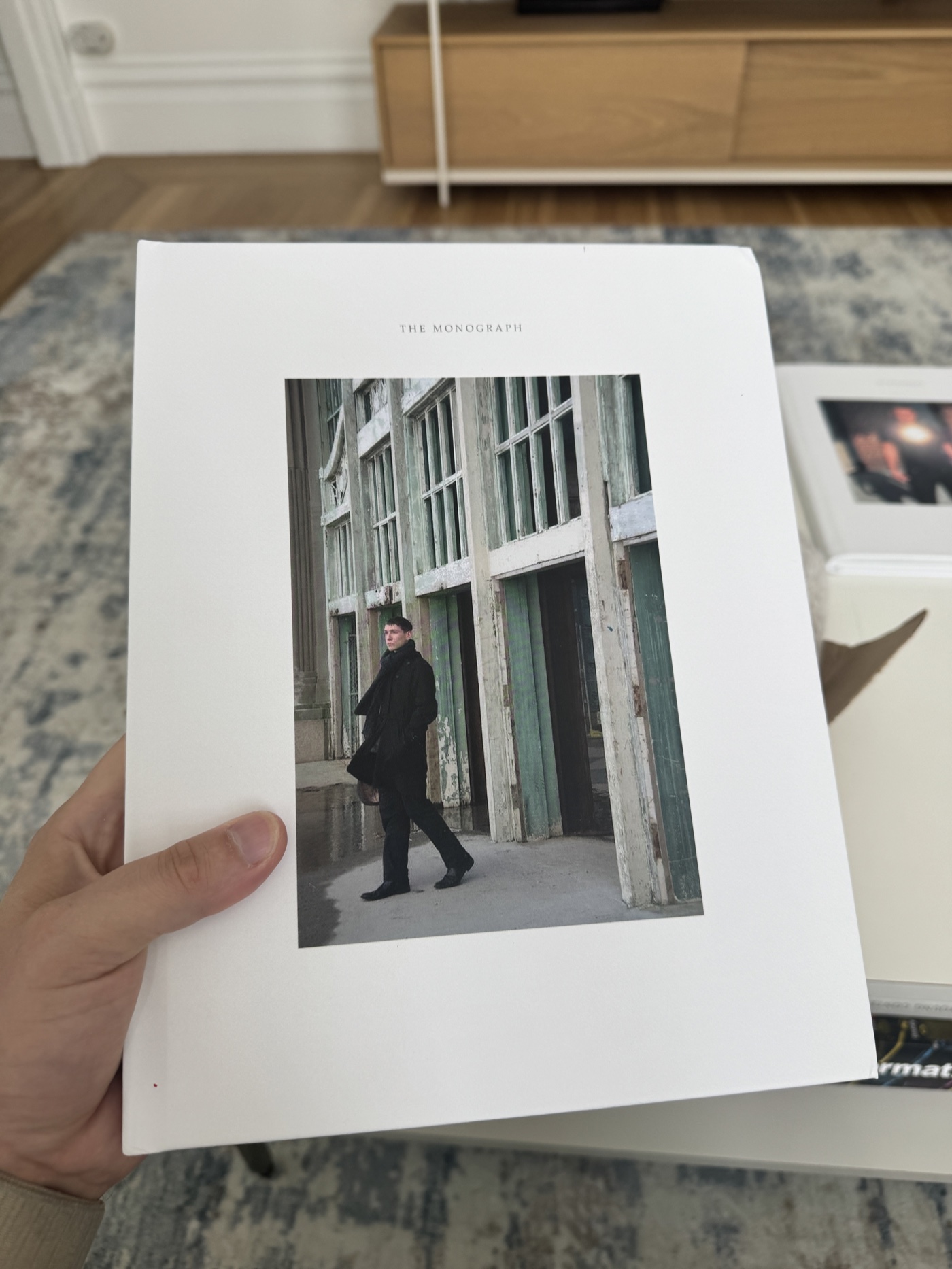 A finished Monograph held in one hand: white hardcover, THE MONOGRAPH stamped above a plate of a figure walking past a row of weathered green doors.